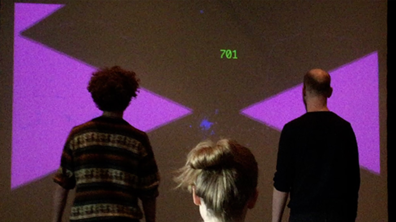 Two people playing closer with one observer standing between them. They standing in front of pink triangles with a blue cluster between them.