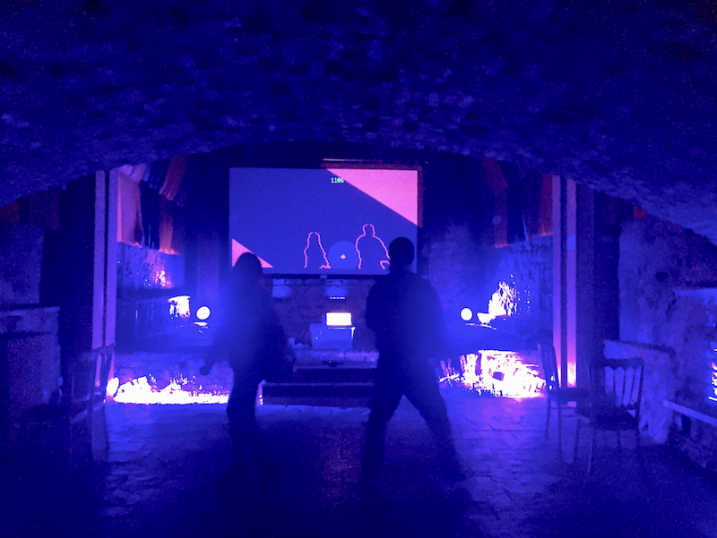 Two players facing a projected screen showing their shared sphere, lit blue in a vaulted stone room