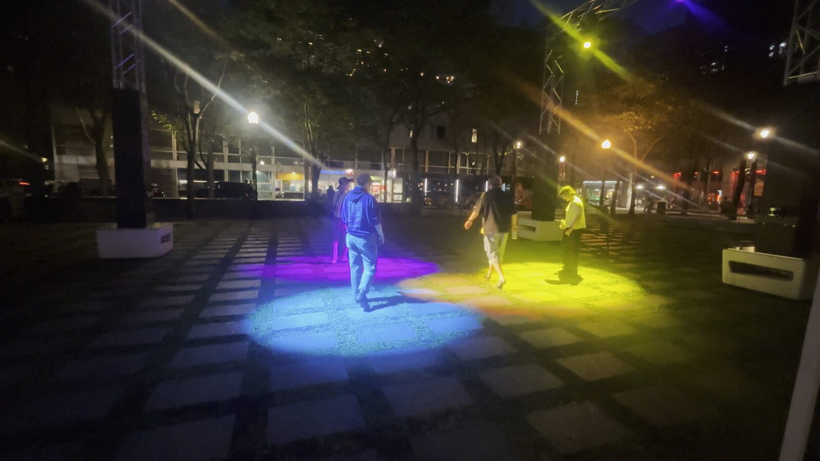 Deux flaques de lumière, l'une bleue, l'autre jaune, abritent chacune une personne sur les dalles du parc.