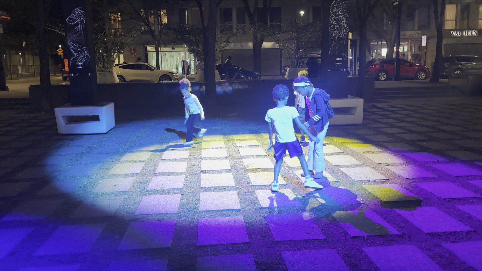 Des gens traversent un damier de lumières arc-en-ciel projetées sur les dalles du parc.