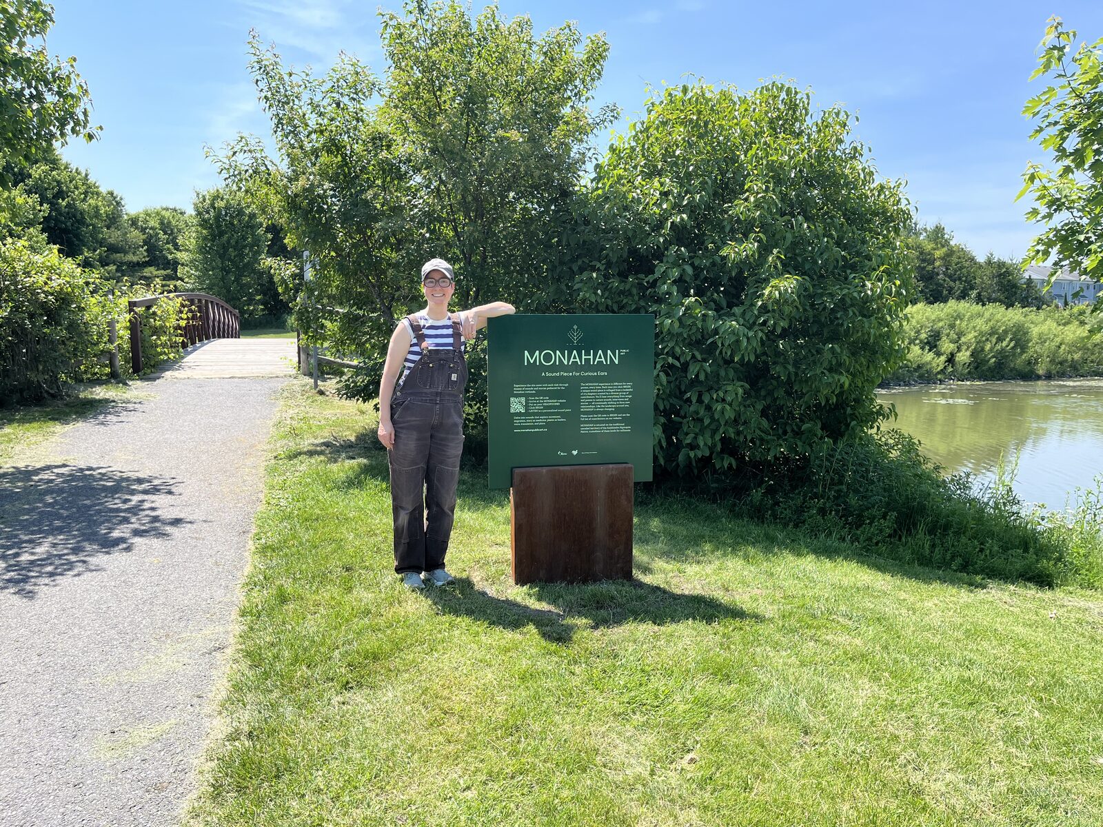 Une personne, sur le sentier au bord de l'eau, montre du doigt le panneau MONAHAN sous un ciel bleu.