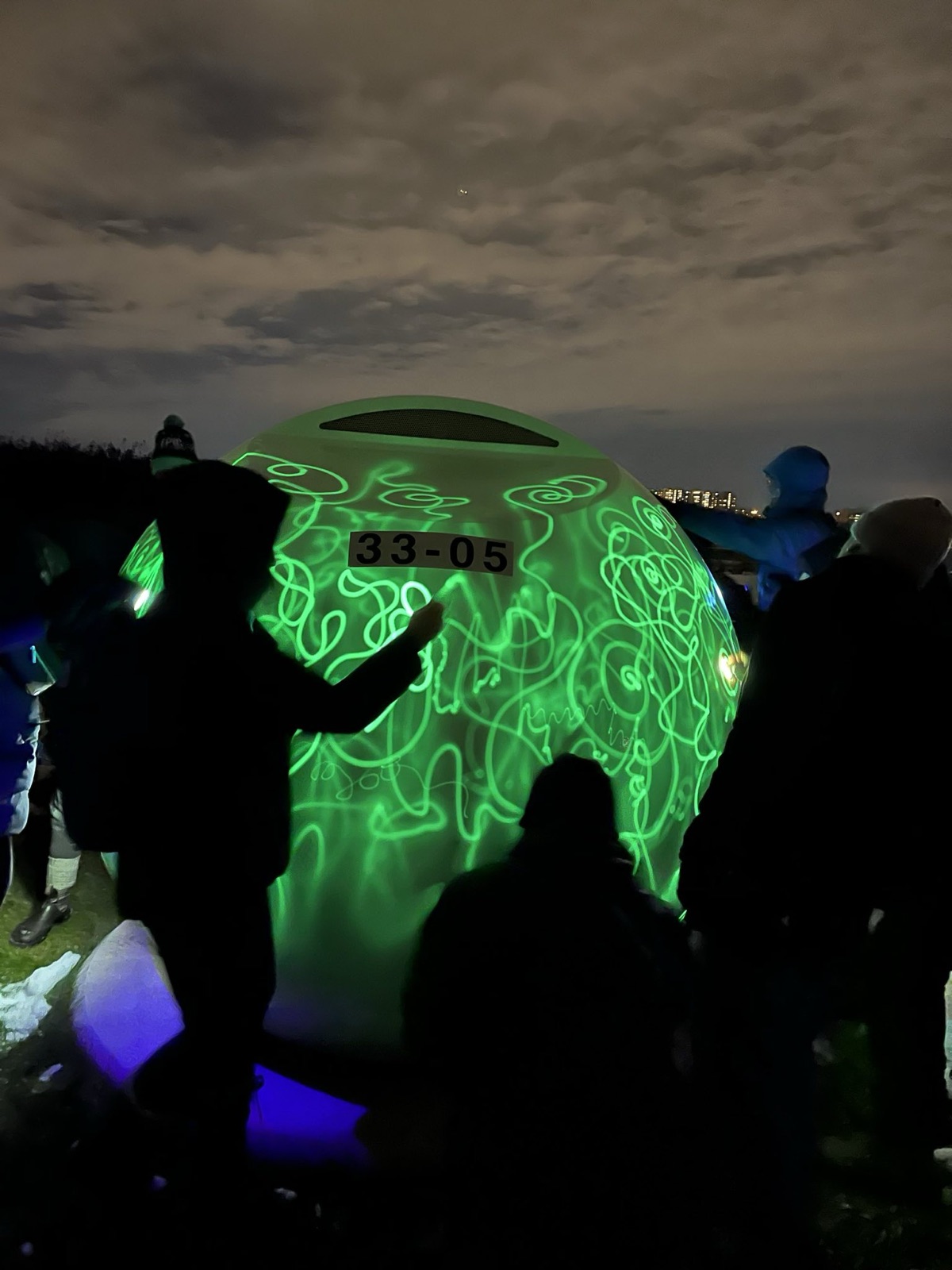 Des silhouettes entourent une orbe qui brille en vert vif, couverte de gribouillis lumineux ; une main la touche.