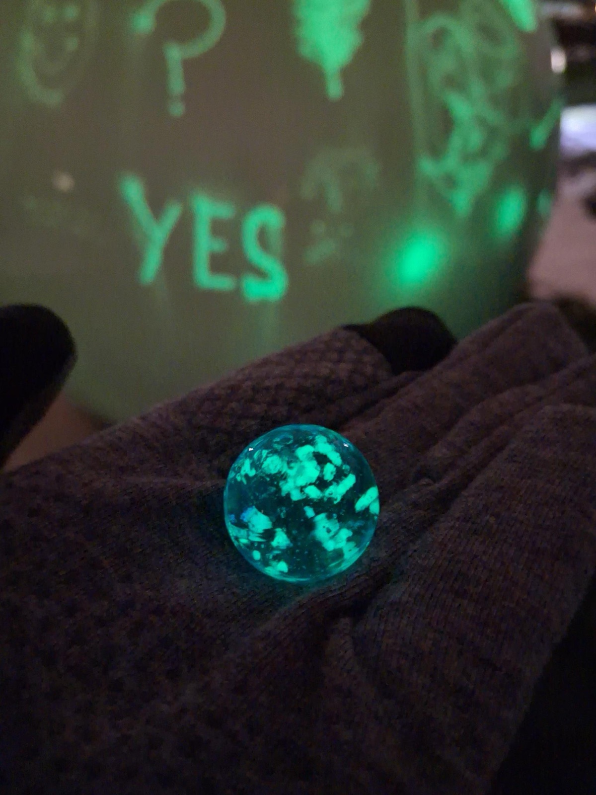 Une bille qui brille en bleu-vert, tenue dans une main gantée, avec le mot « YES » lumineux à l'arrière-plan.