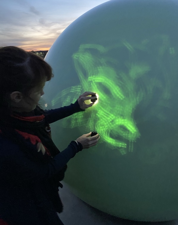 Une personne dessine sur une orbe lumineuse avec une lampe UV, laissant une traînée de lumière verte sur sa surface, au coucher du soleil.