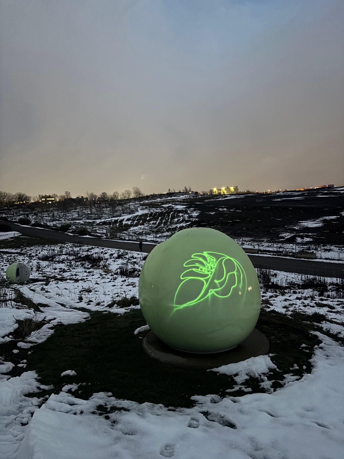 Une orbe vert pâle avec un dessin de feuille tracé en lumière, à l'heure bleue, avec une autre orbe et des bâtiments éclairés au loin.