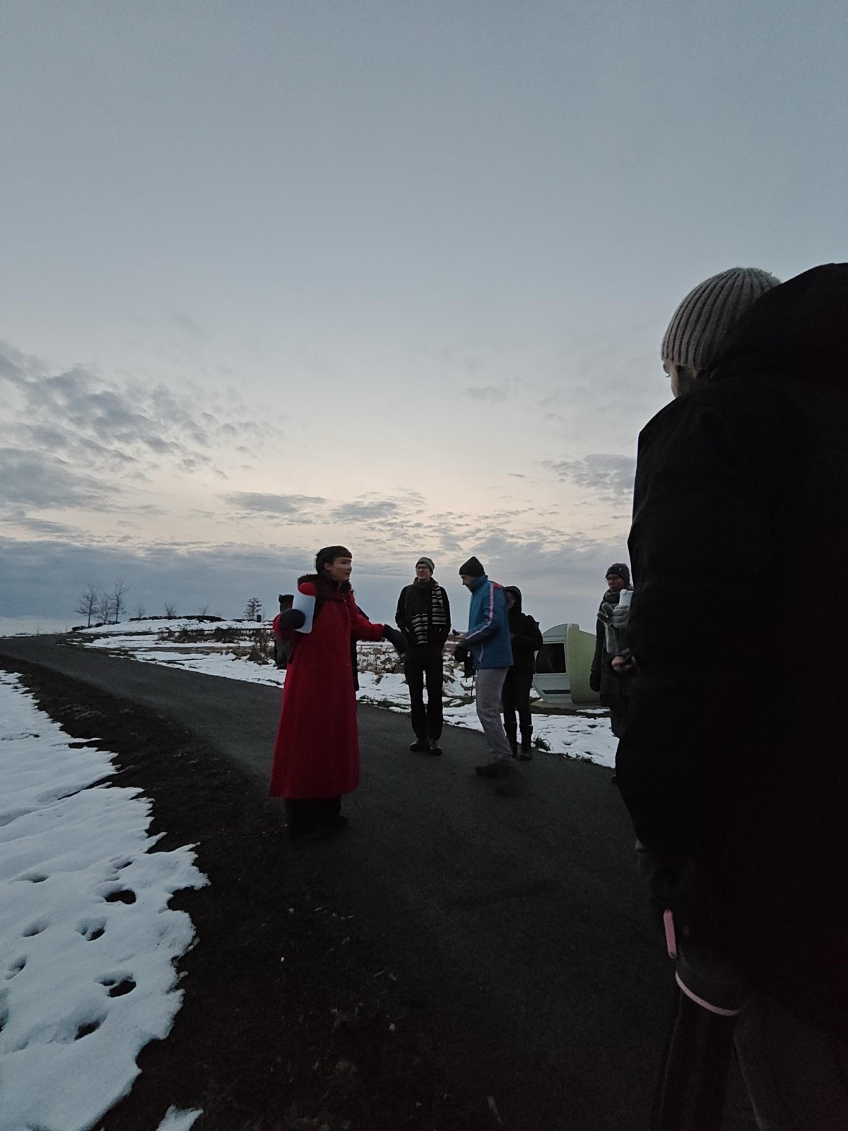 Une personne en long manteau rouge parle à un petit groupe rassemblé sur un sentier enneigé, au crépuscule.