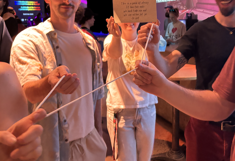 Three people playing the string game together in a crowd, one holding up the rules card