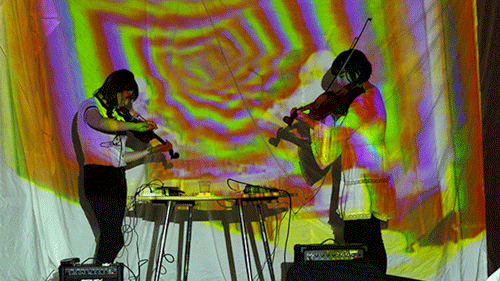 Two people playing violins in front of a multicolour sheet
